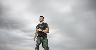 A muscular man running on sandy terrain under cloudy skies, promoting fitness and adventure.