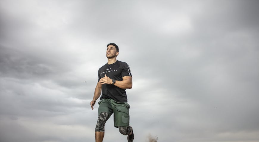 A muscular man running on sandy terrain under cloudy skies, promoting fitness and adventure.